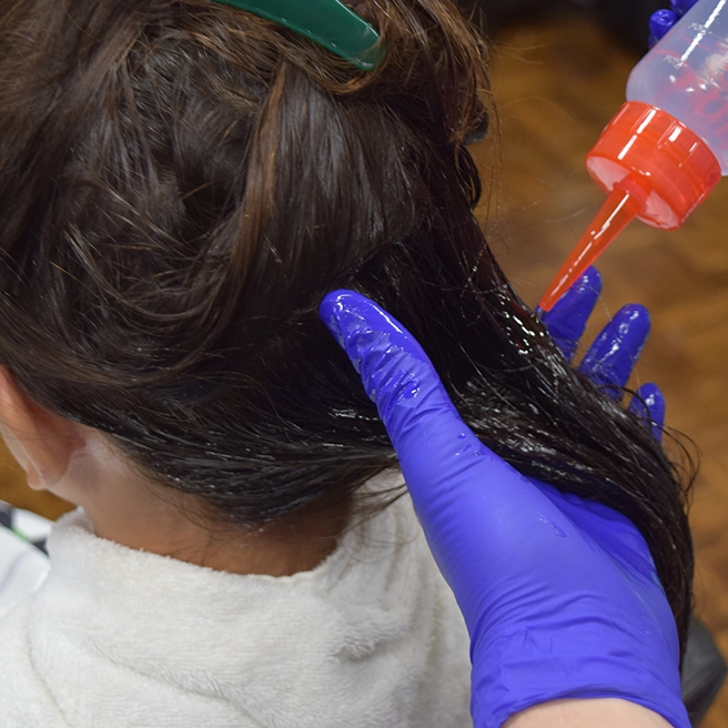 The back of a girls head with brown hair, hands with purple gloves applying oil to the hair.