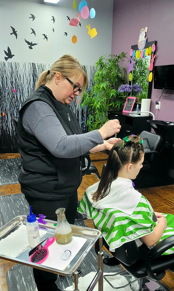Jamie from Jamie's Lice Angels standing behind a young girl in a salon chair. Jamie is combing head lice from the girls hair.